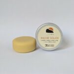 baume solide de couleur jaune clair. à côté se trouve sa boîte en aluminium avec étiquette dans les tons crème, orangé et marron ; logo futur simple