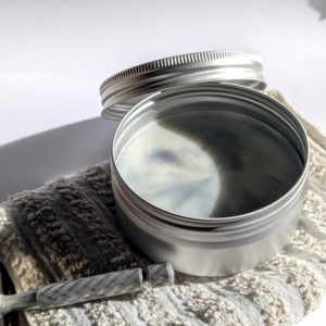 savon de rasage dans sa boîte aluminium, ouverte, posée sur une petite serviette écrue. on aperçoit la manche d'un rasoir.
