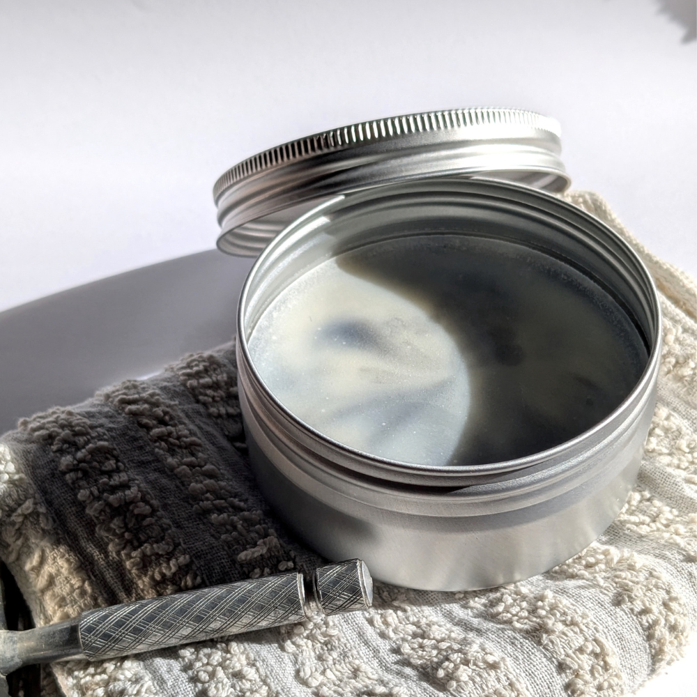 savon de rasage dans sa boîte aluminium, ouverte, posée sur une petite serviette écrue. on aperçoit la manche d'un rasoir.